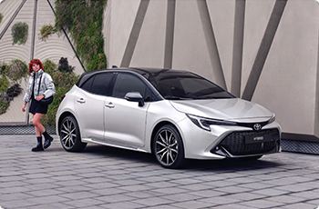 Stříbrná Toyota Corolla hatchback s černou střechou zaparkovaná před moderní budovou; vedle ní jde žena.